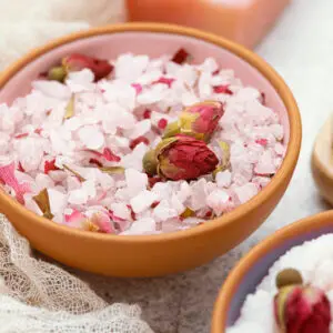 Wooden bowl with pink scrubbing salts in it, scattered with flower petals