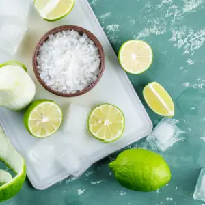 Lemons with salt, ice cubes on plaster and cutting board background, flat lay.