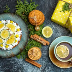 hand-holding-cup-black-tea-creamy-delicious-cake-fir-branches-lemon-cinnamon-limes-yellow-gift-boxes-dark-background
